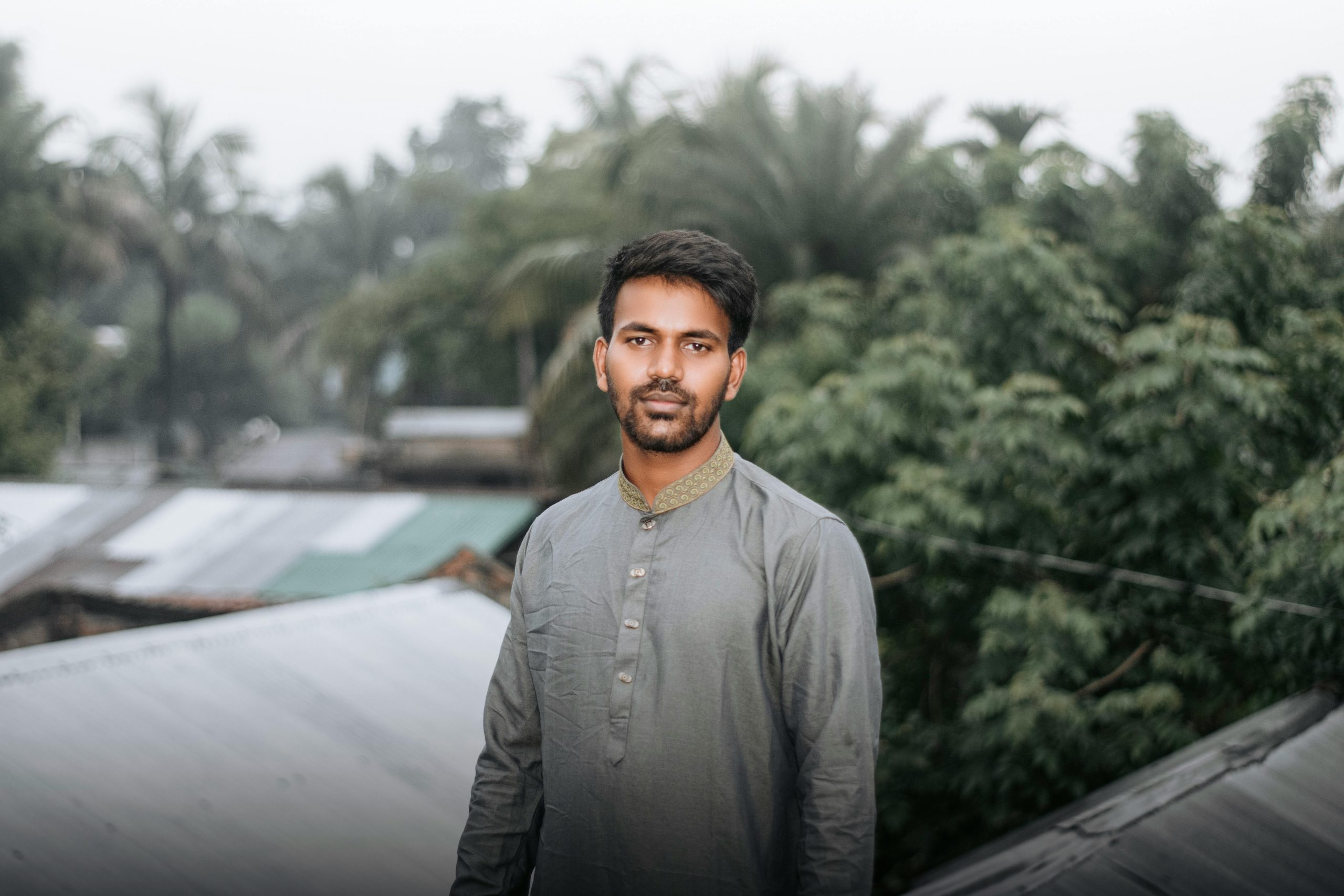 A young South Asian man stands outdoors with lush greenery in the background, exuding calmness.